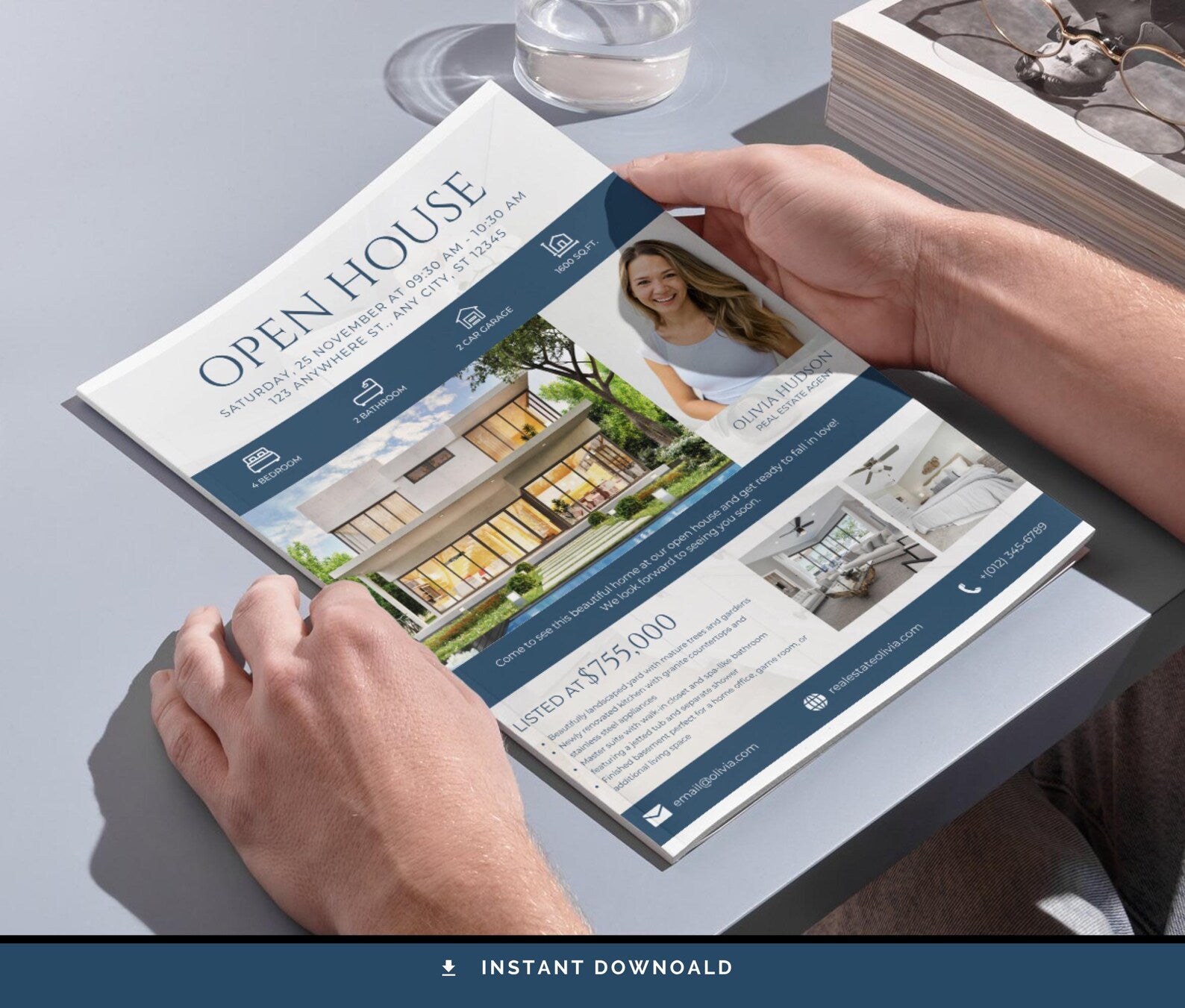 A person holds an open real estate open house brochure over a gray tabletop. The brochure has a white and navy design with property photos, text, and a woman's portrait.
